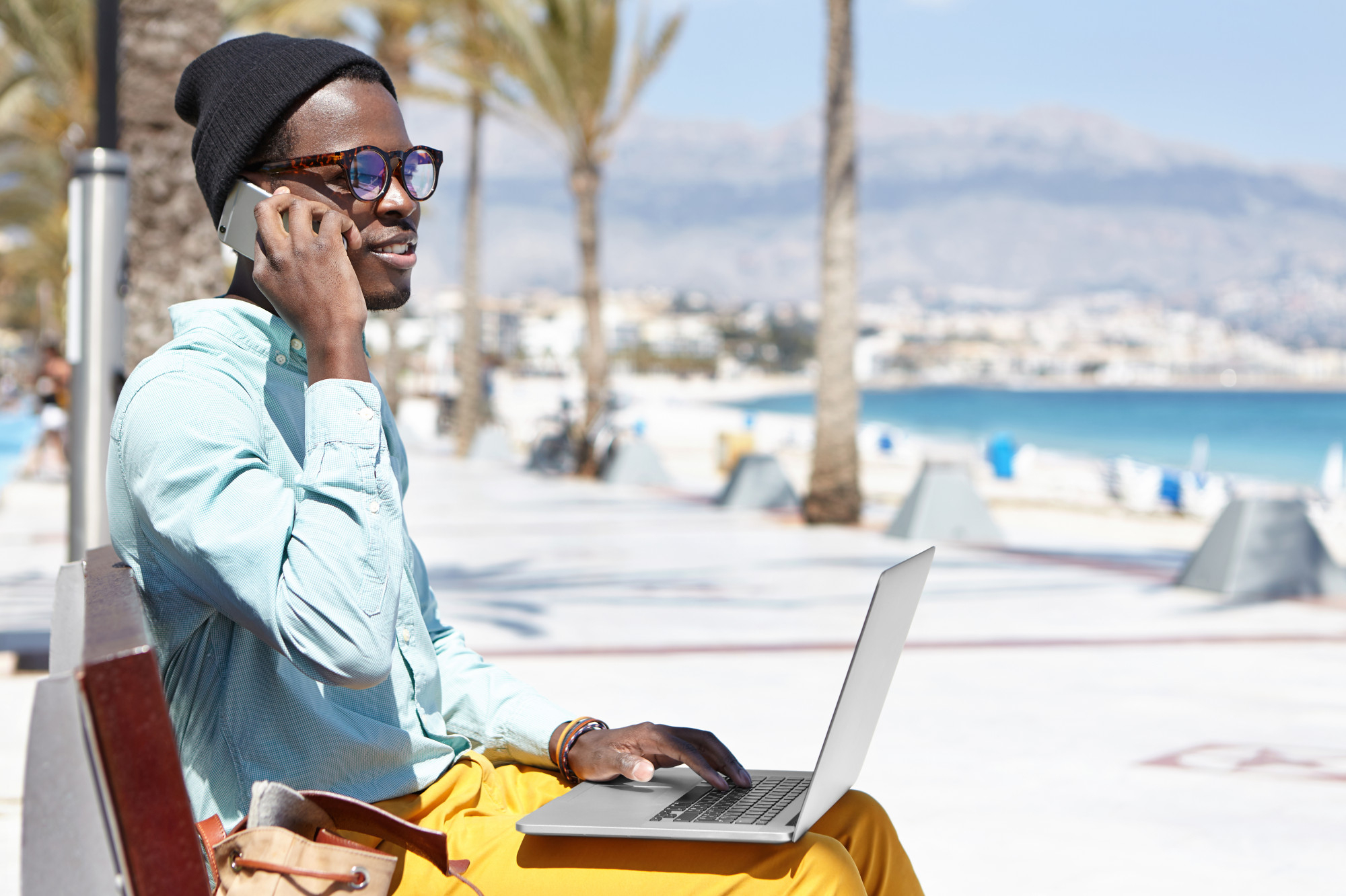 <p>A man sitting on a boardwalk working remotely with his laptop in his lap while talking on his mobile.</p>