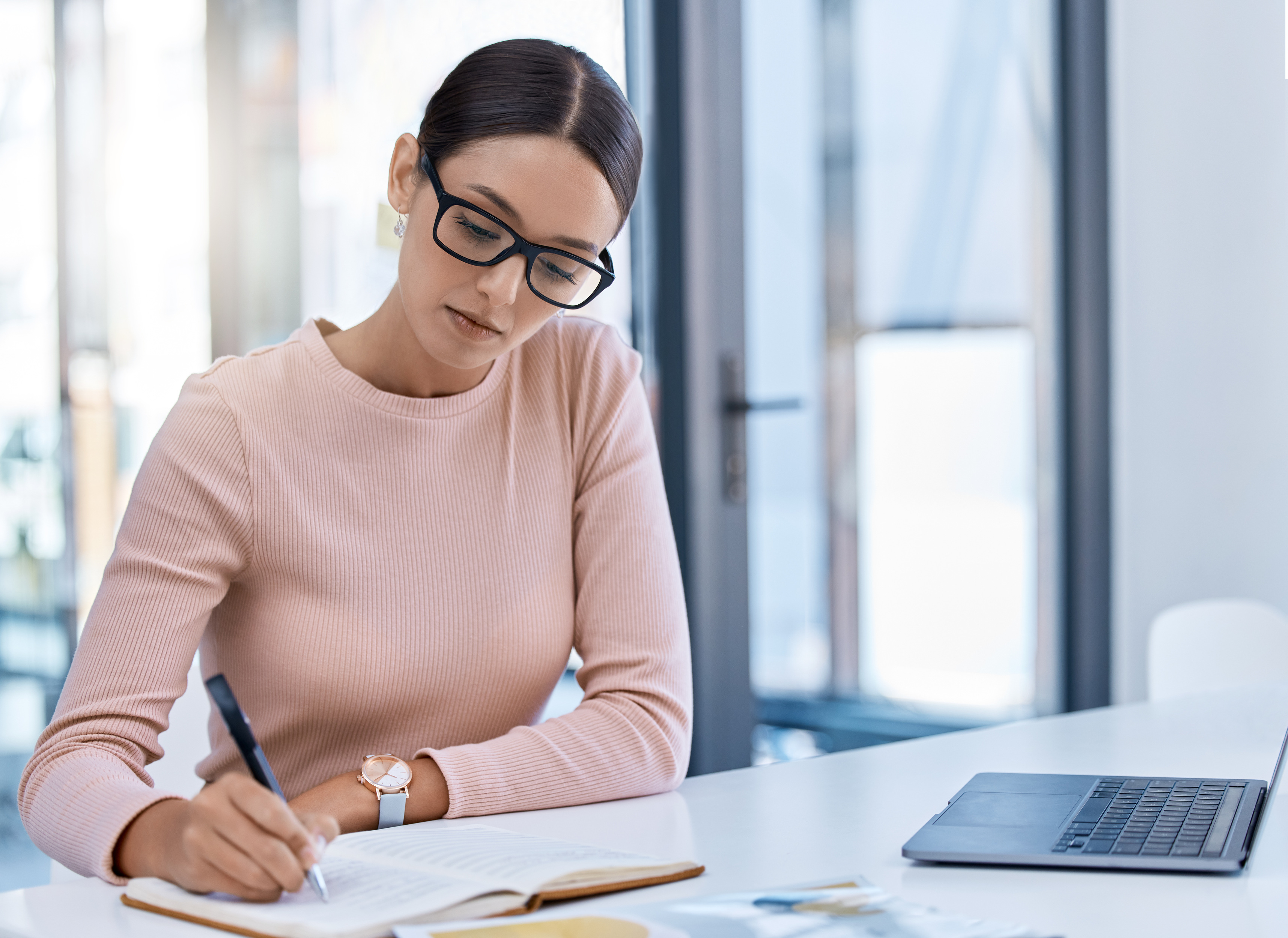 <p>A woman with glasses writing down notes while working on a laptop in the office</p>