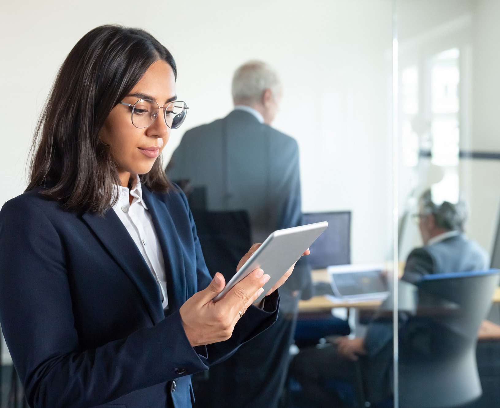 <p>Smart-looking woman with glasses looking at a tablet, while two male colleagues are talking in the background.</p>