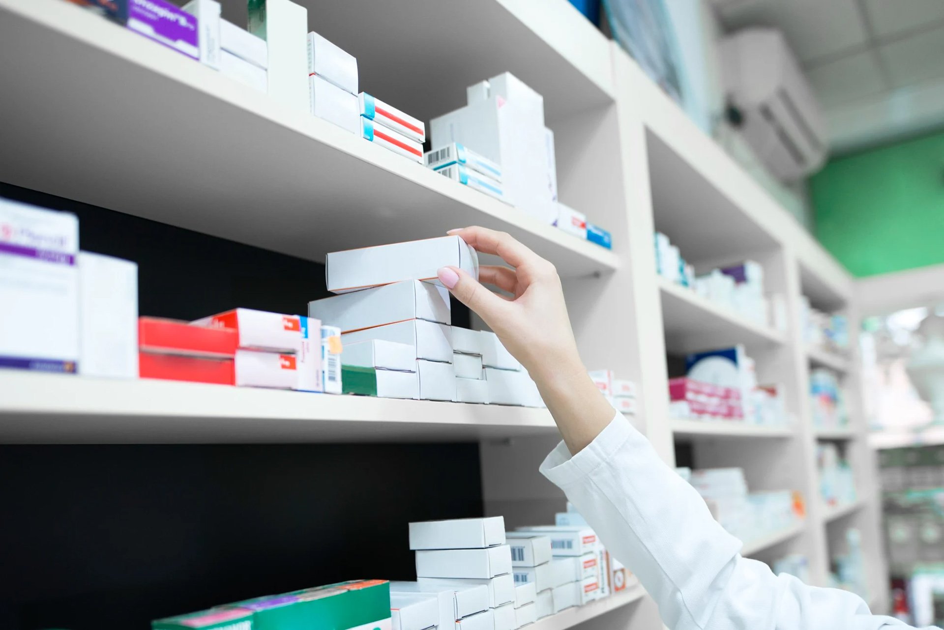 Pharmacist's hand reaching for a medicine box on a shelf filled with various pharmaceutical packages.