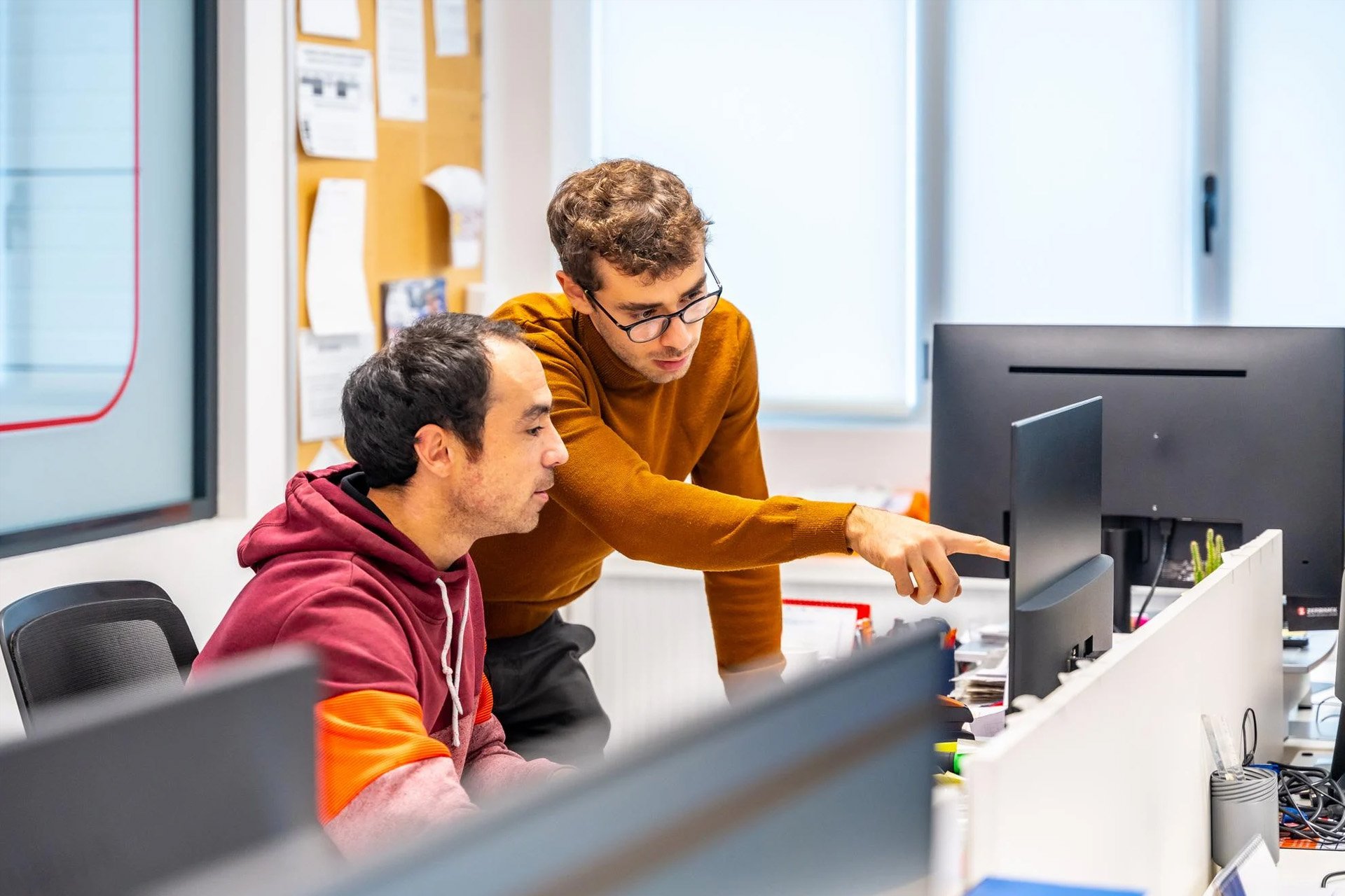Two men working together at a computer in a bright office, one pointing at the monitor.