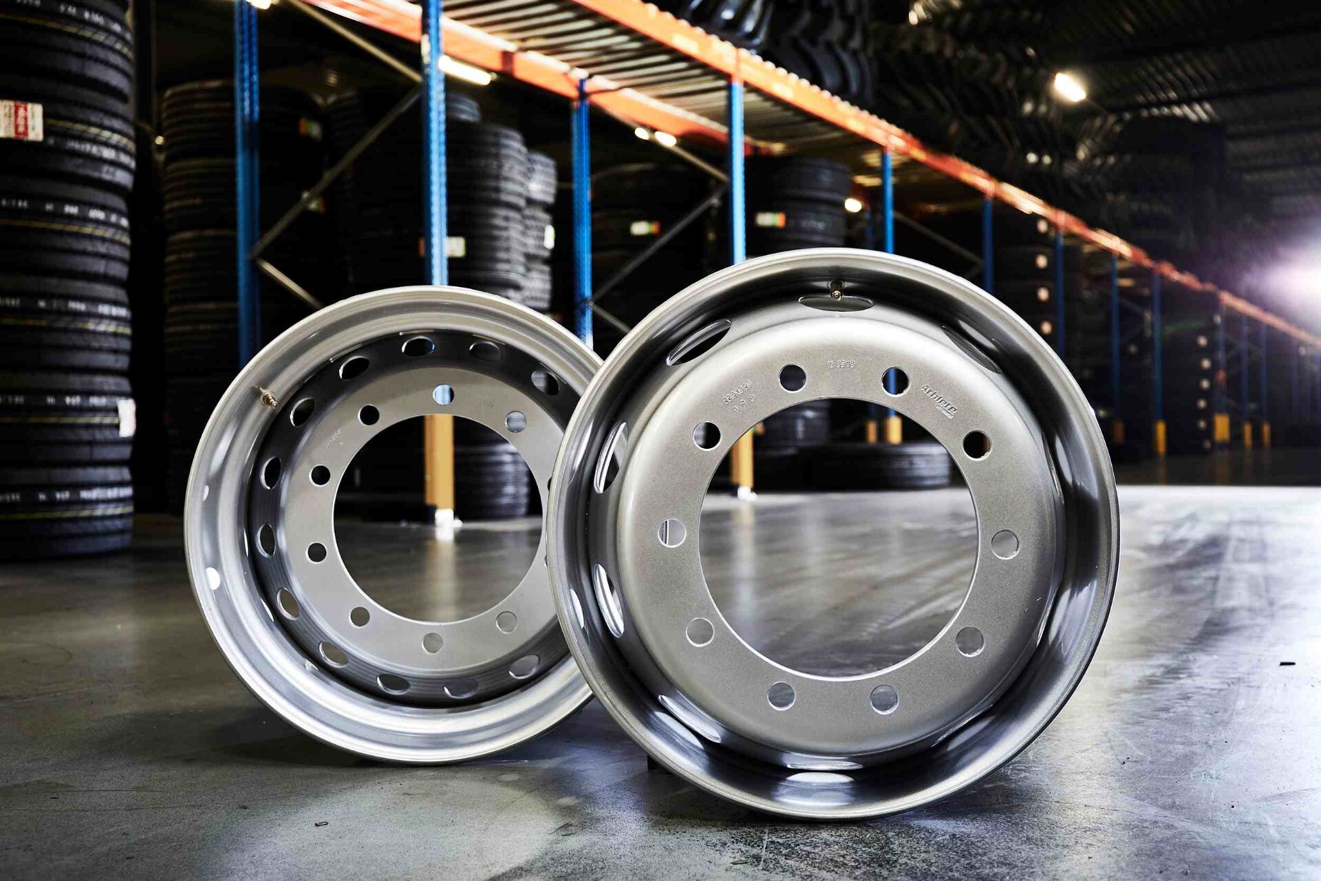 Two silver wheel rims placed on the floor of a warehouse with stacks of tires in the background.