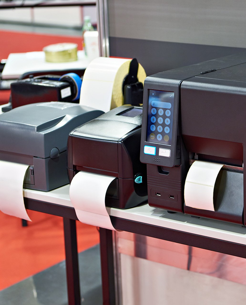 Close-up of several label printers on a table, printing labels on white paper, with one printer displaying a digital control panel.