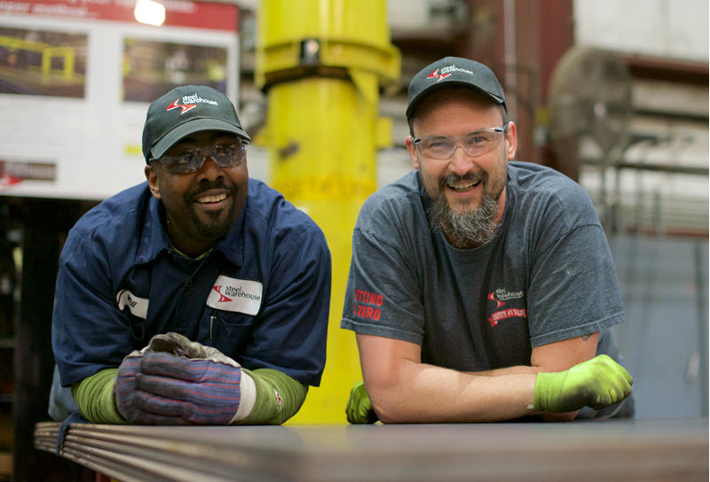 Two factory workers leaning on a metal surface and smiling inside a manufacturing facility.