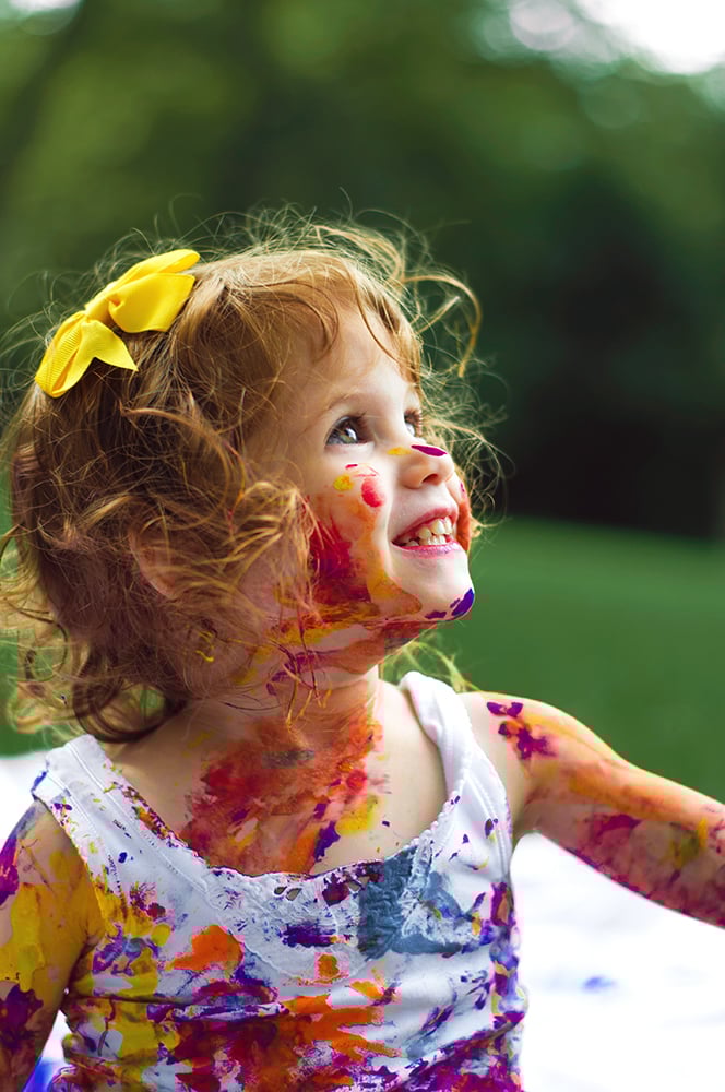 Young girl with a yellow bow smiling and covered in colorful paint.