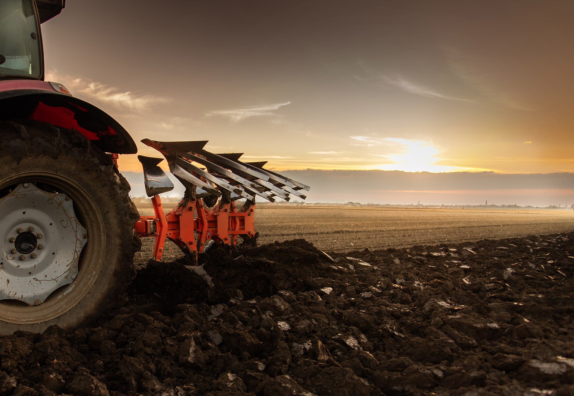 A tractor plowing a field at sunset, with gleaming metal plowshares and freshly turned dark soil. Soft clouds illuminated by the sun.