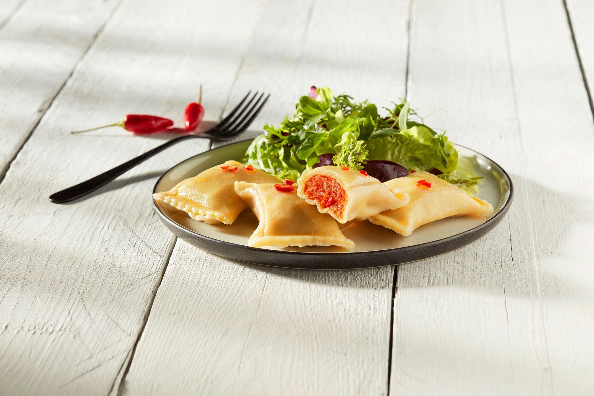 Plate of stuffed pasta with red filling, garnished with chili slices and served with fresh green salad on a white wooden table.