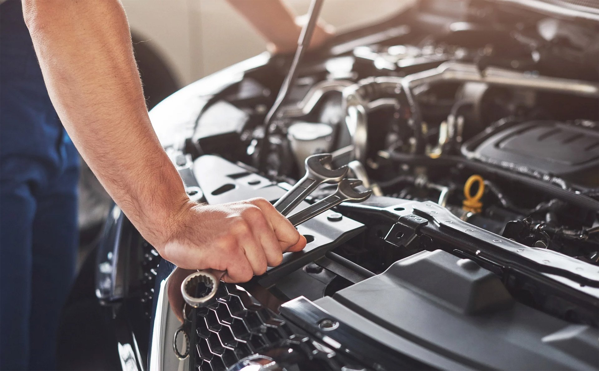 Mechanic holding wrenches while working on a car engine in an auto repair shop.