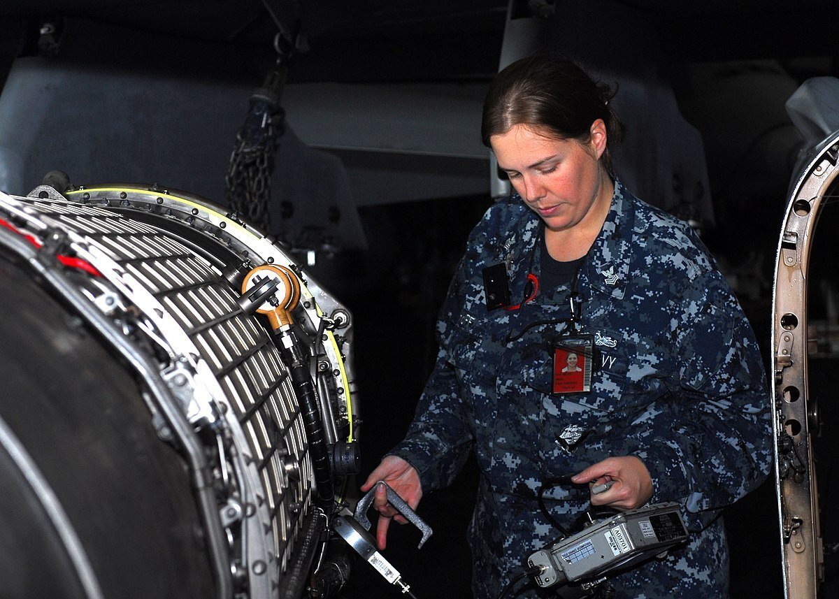 A technician in a navy uniform inspects a jet engine, using tools to perform maintenance in a dimly lit environment.