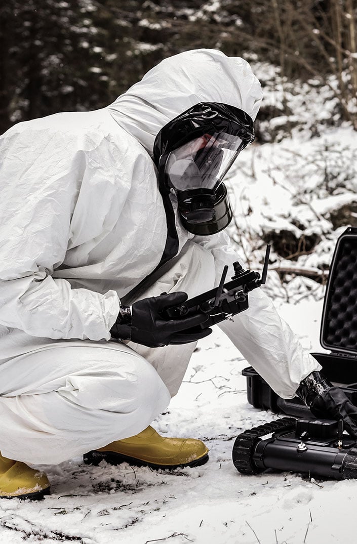 A person in a white hazmat suit and gas mask checks equipment in a snowy forest, crouching beside a black case and robotic device.