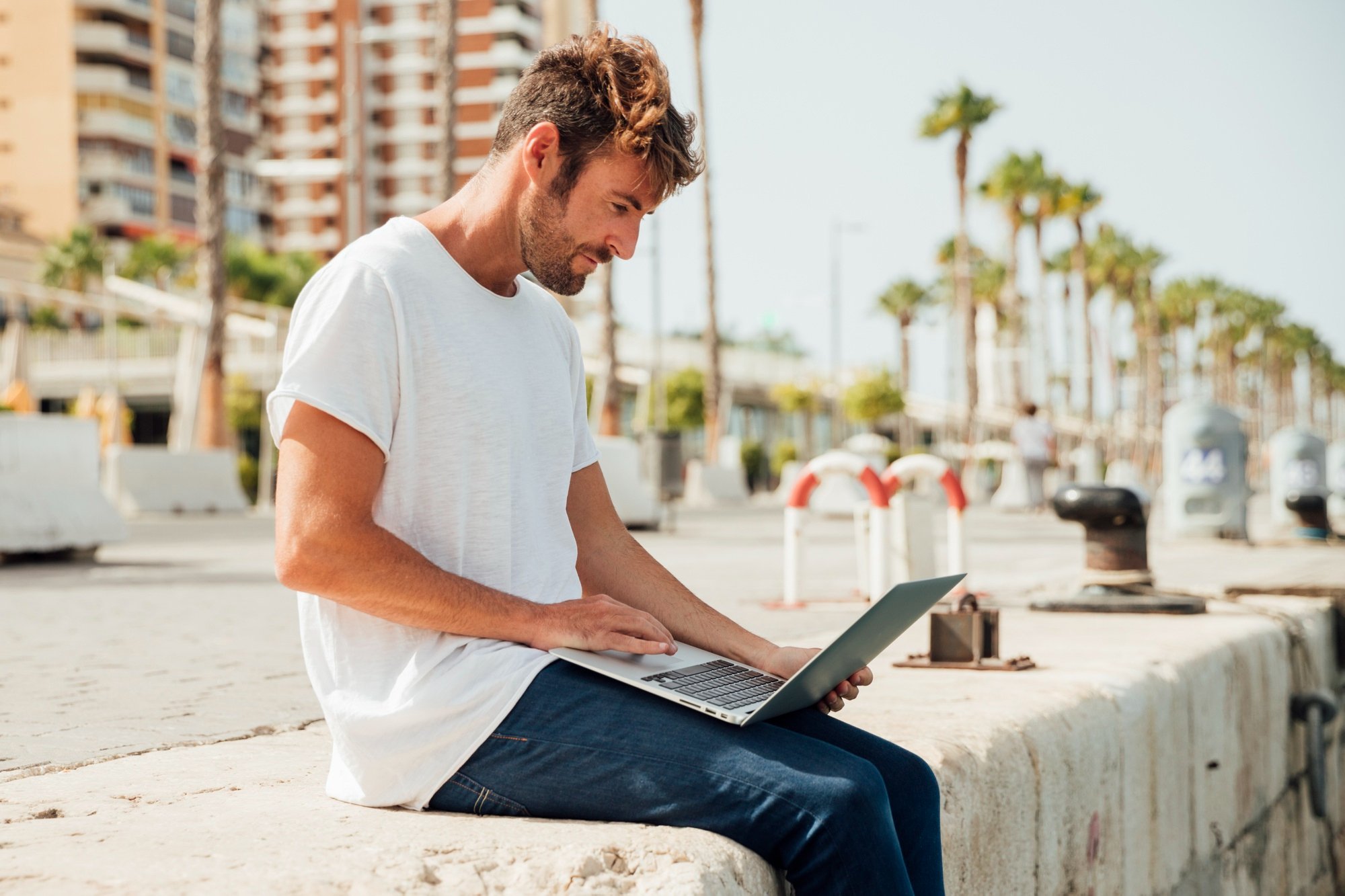 Man sitting on a dock using a laptop with palm trees and buildings in the background.
