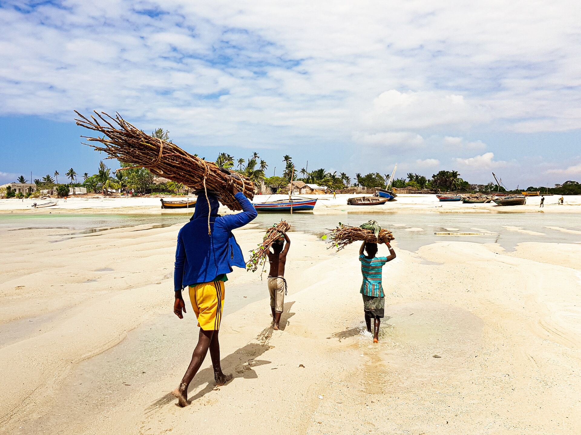 Three people carrying bundles of sticks on a sandy beach near fishing boats.