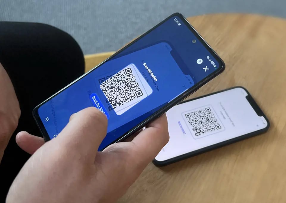 A person holds a smartphone displaying a QR code, while another phone in the background shows a different QR code on its screen.