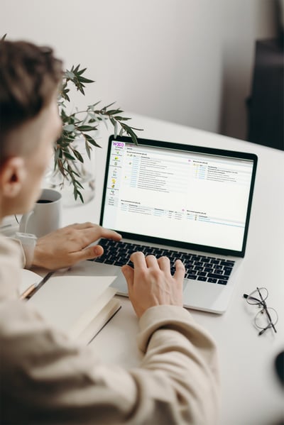 En person använder en bärbar dator vid ett vitt skrivbord, fokuserad på startsidan av W3D3 från Formpipe med olika organisatoriska verktyg och med en anteckningsbok bredvid.
