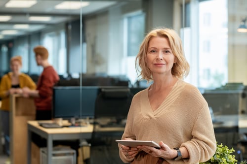 mature-blond-businesswoman-casualwear-standing-large-modern-office-using-tablet-against-two-young-colleagues