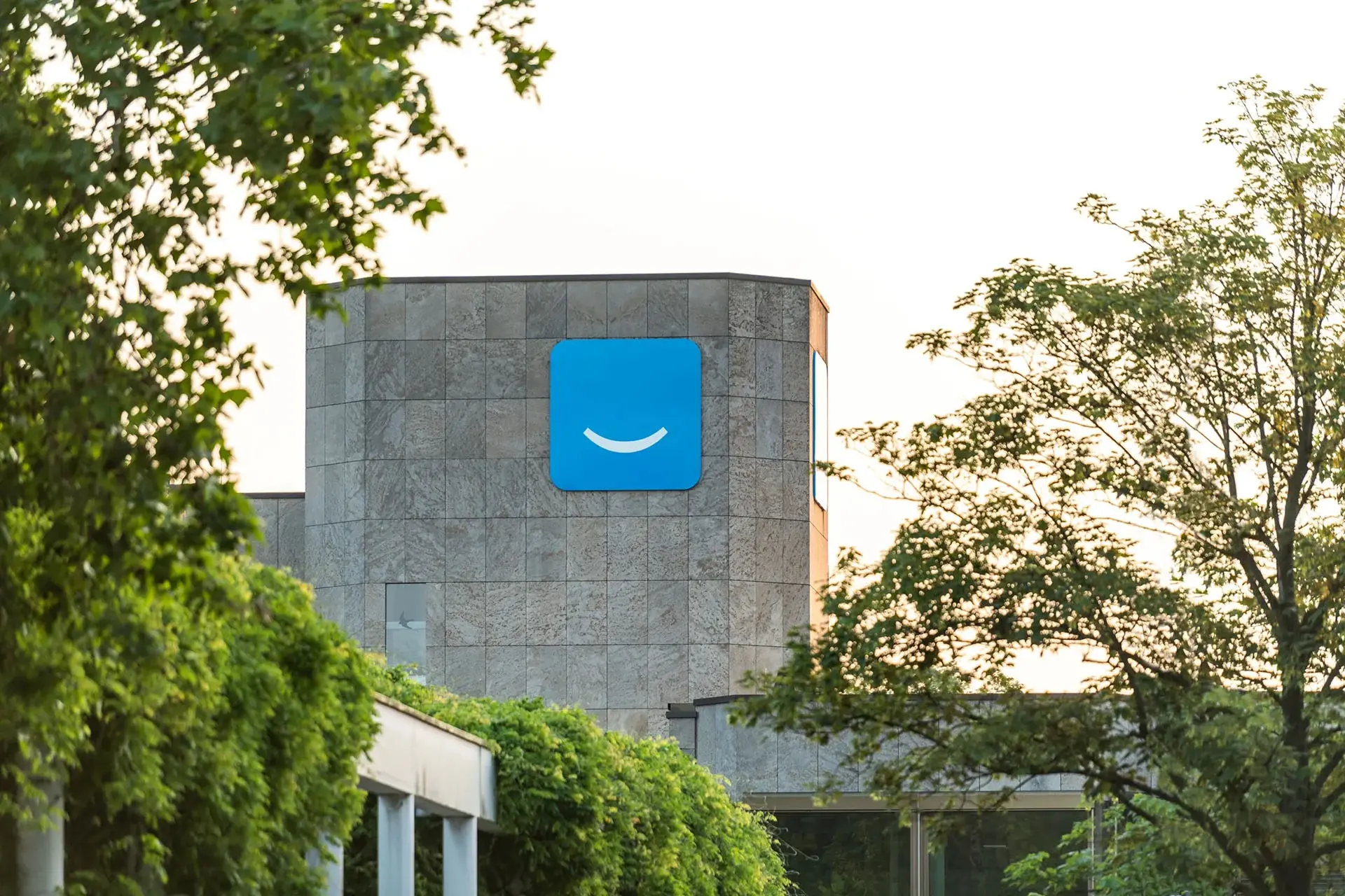 Modern building exterior with a large blue logo, featuring a white smile, on a stone wall. Framed by lush green trees under a clear sky.