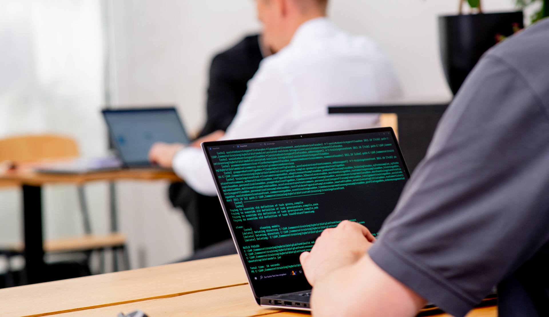 A person is typing on a laptop displaying green coding text, with another individual working on a laptop in the background. A person is typing on a laptop displaying green coding text, with another individual working on a laptop in the background.