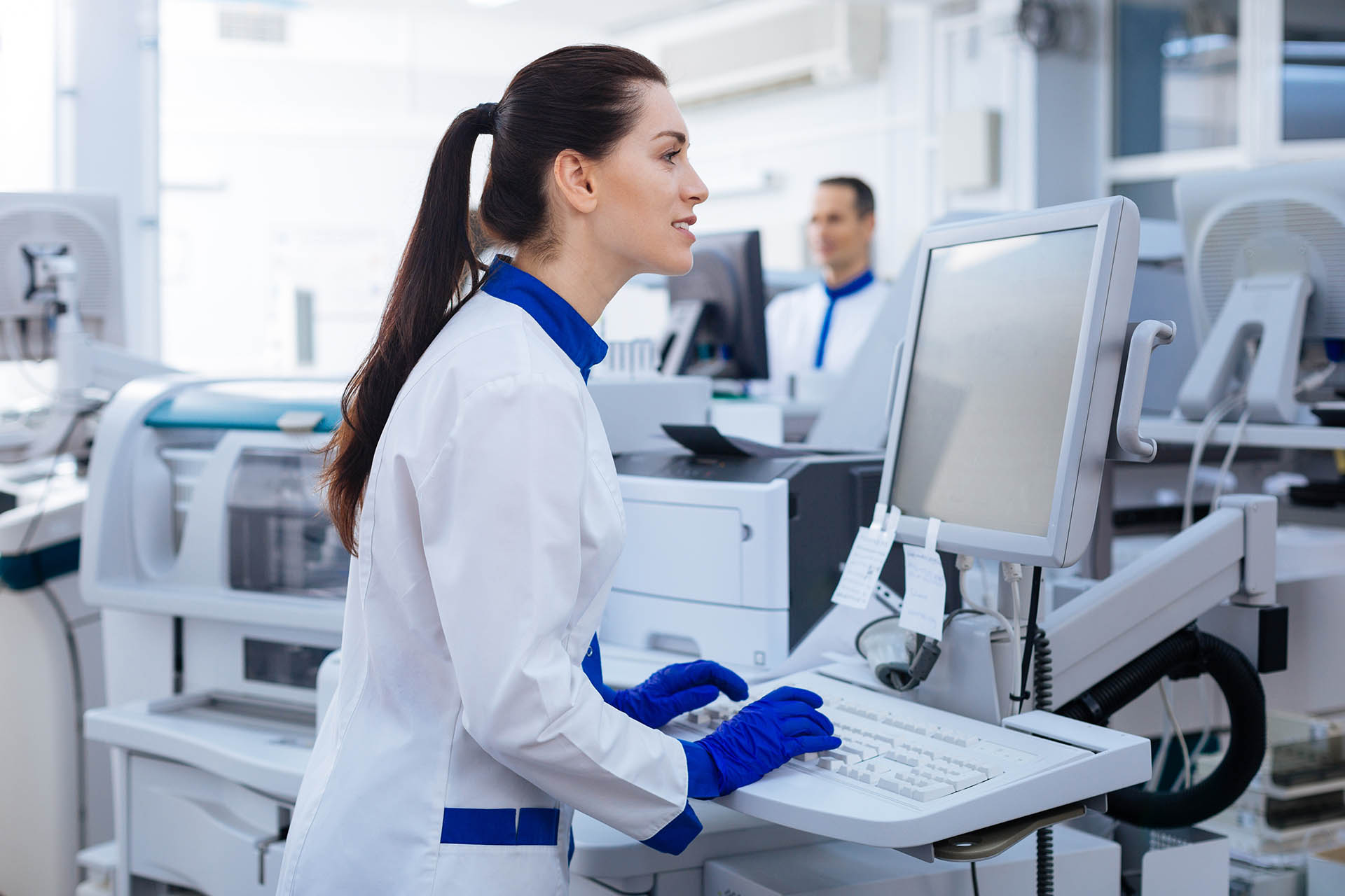 A laboratory technician in a white coat and blue gloves works at a computer, surrounded by medical equipment in a bright workspace. A laboratory technician in a white coat and blue gloves works at a computer, surrounded by medical equipment in a bright workspace.